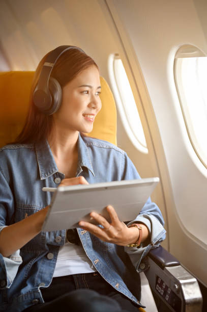 Charming asian woman passenger at window seat with headphones using portable tablet Charming millennial asian woman passenger at her window seat with her wireless headphones using portable tablet and looking out the plane's window. economy class stock pictures, royalty-free photos & images