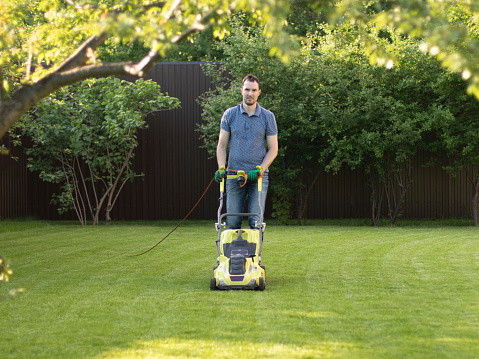 A young man cuts a lawn by lawn mower. Summer work in the garden