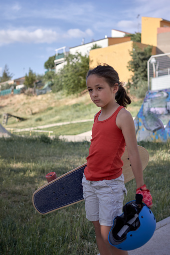 girl in shorts and red tank top with a skateboard under her arm and a blue helmet in her left hand has just arrived at the skatepark in her town.