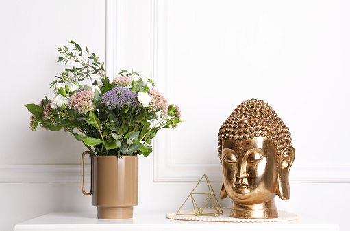 Stylish ceramic vase with beautiful flowers and golden Buddha sculpture on table near white wall
