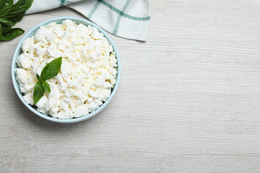 Fresh cottage cheese with basil in bowl on light wooden table, flat lay. Space for text
