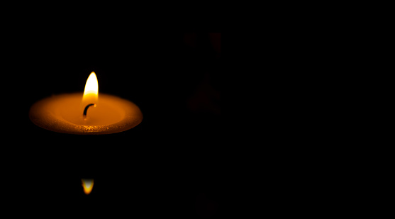 Light candle floating on water with a reflection of flame and bright orange glow set against a black background.