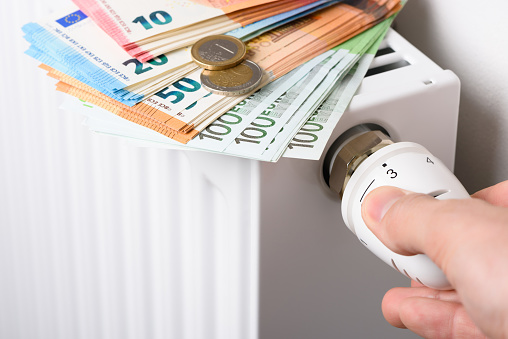 Hand adjusting the valve knob of heating room radiator temperature thermostat with stack of euro money banknotes and coins on it. Expensive home heating costs and rising energy bill prices for winter cold season