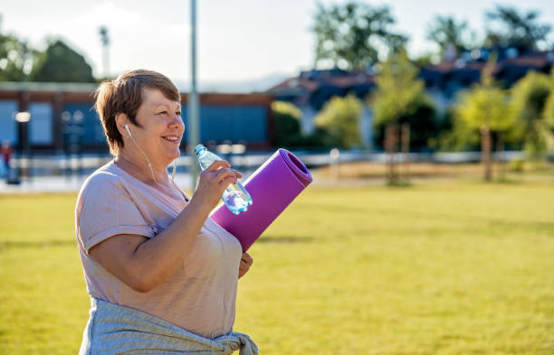 happy smiling senior plus size woman with earphones holding yoga mat outdoors drinking water resting after exercises at warm sunny summer day - plussize model fotos stockfoto's en -beelden