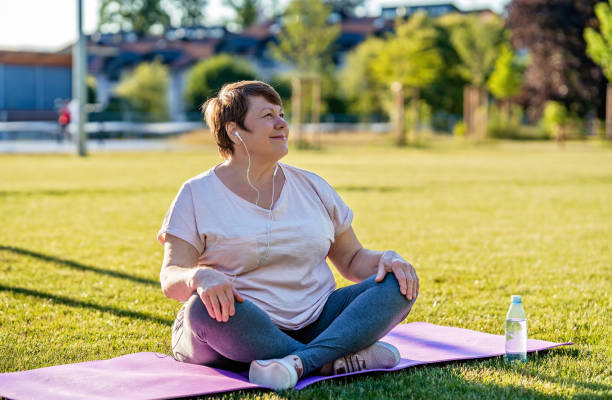 Relaxed senior plus size woman with earphones sitting on yoga mat on green grass outdoors resting after exercises at warm sunny summer day stock photo
