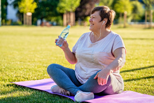Relaxed senior plus size woman with earphones sitting on yoga mat on green grass outdoors resting after exercises drinking water at warm sunny summer day stock photo