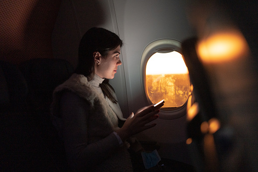 Woman traveling by plane and looking through window and using mobile phone