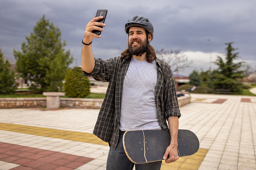 Urban young Caucasian man, taking selfie with mobile phone, while taking a break from skateboarding in the public park