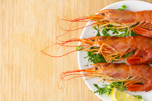 Three whole jumbo shrimp on a herb salad with fresh lemon, on a light wooden background.  Shot from above with copy space to the left of the image. Stock photo.