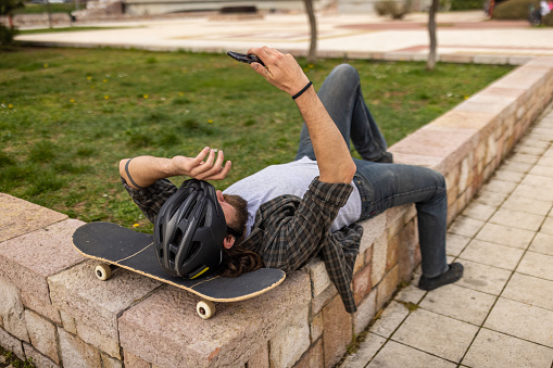 Urban young Caucasian man, taking selfie with mobile phone, while taking a break from skateboarding in the public park