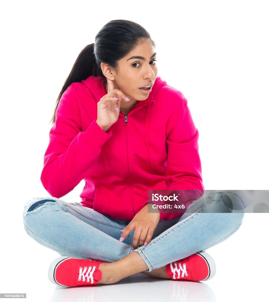 Generation z young women sitting on floor in front of white background wearing hooded shirt Front view of aged 20-29 years old who is beautiful with black hair generation z young women sitting on floor in front of white background wearing hooded shirt who is curious who is whispering Deafness Stock Photo