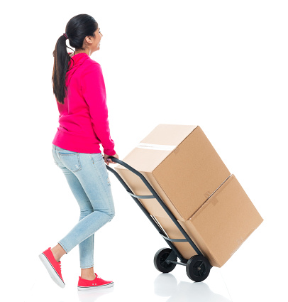 Rear view of aged 20-29 years old who is beautiful with black hair generation z young women manual worker walking in front of white background wearing pants who is relocation and holding cardboard box and using hand truck