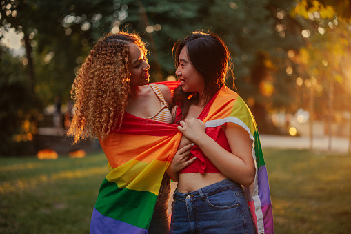 A young female multiracial couple is outdoors with a rainbow flag around them.