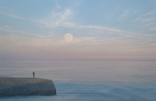 A large moon rises over the sea, being wached by a person on the Cobb in Lyme Regis