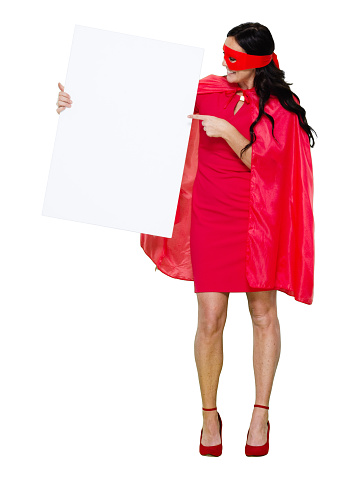 Front view of aged 40-44 years old who is beautiful with black hair caucasian female hero standing in front of white background wearing mask - disguise who is happy who is pointing and holding banner sign with copy space