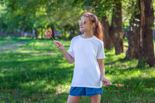 A teenage girl in a white casual T-shirt with multi-colored lollipops in her hands. Front view of t-shirt mockup for design print. Summer vacation in the park. Horizontal photo.