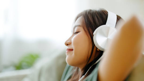 Young asian woman listening to music on couch in living room at home. Happy asia female using mobile smartphone, wearing headset and sitting on sofa stock photo