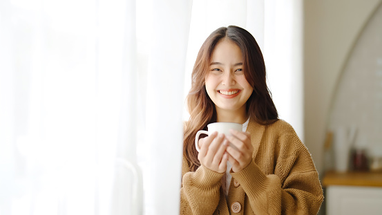 Young asian woman standing beside window and holding mug in living room at home, She drinking milk after wake up in the morning
