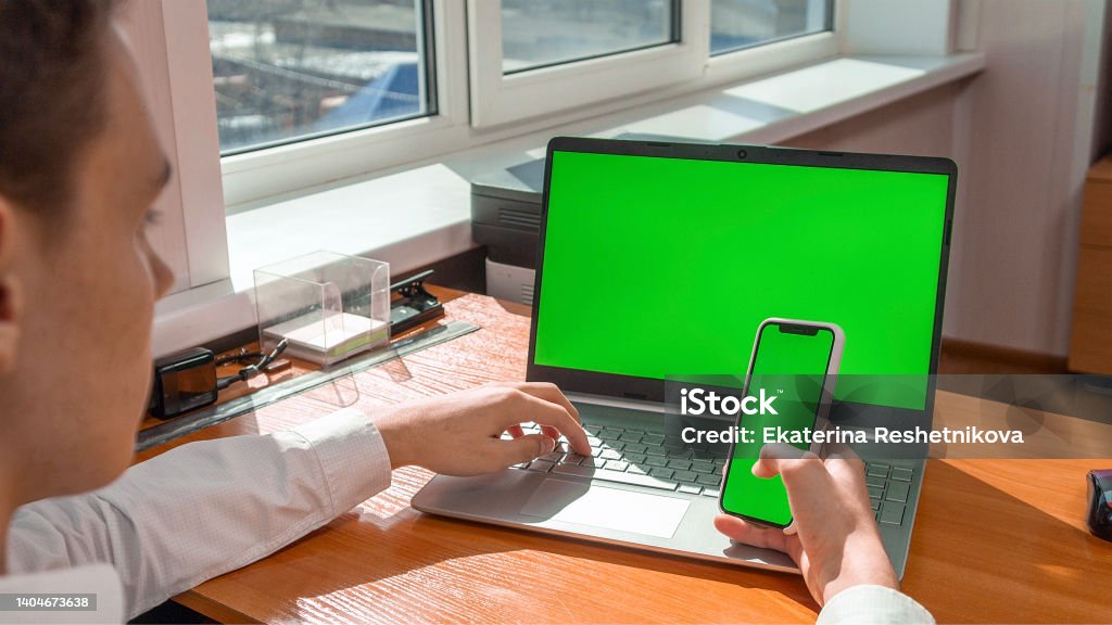 A young man in the office at a laptop with a green screen, holding a phone with a green screen Processed with Lensa with Auto Adjustments Brand Name Smart Phone Stock Photo