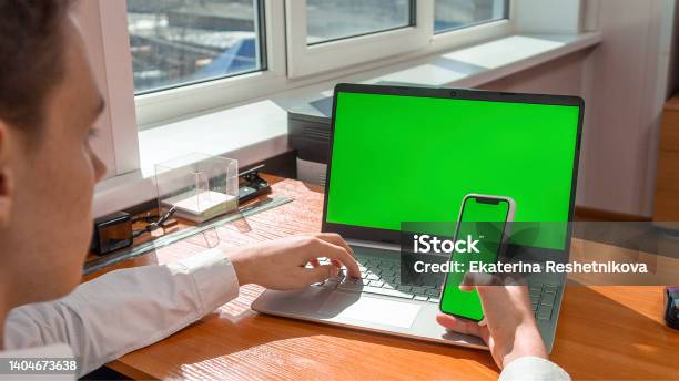 A Young Man In The Office At A Laptop With A Green Screen Holding A Phone With A Green Screen Stock Photo - Download Image Now