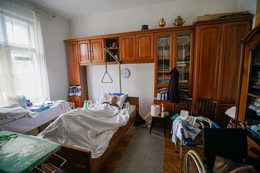 View of Senior, 95 years old immobile woman, alone at home,  lies in her bed in her bedroom. In the room there are the day care supplies like wheelchair, toilet chair, ironing board, napkins everything she needs.