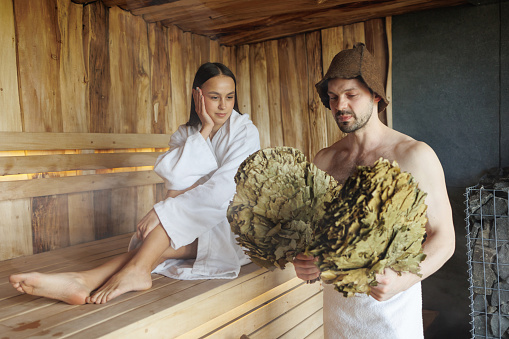 Muscular man with brooms in the sauna and his woman sitting on the background. Beautiful couple in the sauna