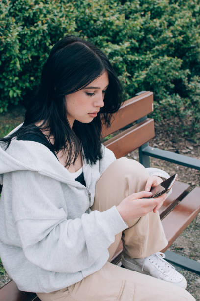 屋外のベンチに一人で座りながらスマートフォンを使用する美しいティーンエイジャーの女の子 - teenager problems typing teenagers only ストックフォトと画像