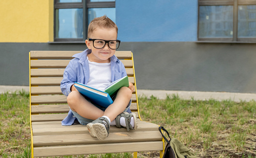 Funny boy with glasses sits and reads a book on the bench. The concept of education.