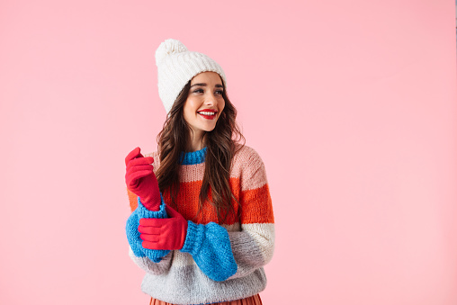 Beautiful young smiling woman wearing sweater and a hat standing isolated over pink background