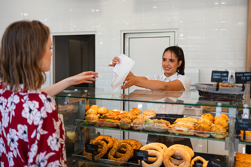 A smiling saleswoman at a bakery gives a paper bag with pastry to a female customer.