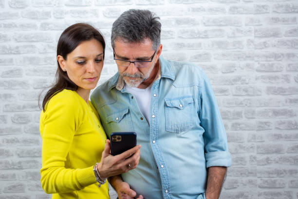 personas felices caucásicas en estilo casual y de diferentes edades sosteniendo el teléfono celular, revisando y usando messenger. hombre y mujer escribiendo en teléfonos móviles aislados en un fondo de pared de ladrillo blanco - couple dependency standing men fotografías e imágenes de stock