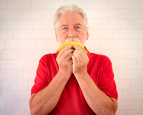 Serious senior man with a white beard and in red polo shirt isolated on a white background holds a banana in front of his mouth