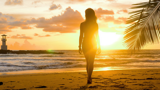 Cinematic video of woman walking on tropical beach on sunset. Female tourist on summer vacation. Slow motion of female traveler walking barefoot by seaside with palm trees