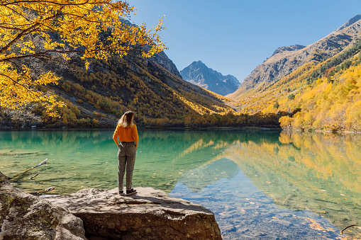 Woman stay on rock and look at crystal lake in the autumnal mountains. Mountain lake and traveller girl