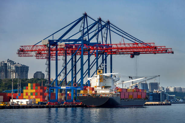 cargo ship moored for loading at container yard with container handling gantry crane or ship-to-shore crane unloading goods from deck to cargo port area, background cityscape with high-rise building. buque de carga amarrado para carga en el patio de contenedores con grúa pórtico de manejo de contenedores o grúa de barco a tierra que descarga mercancías desde la cubierta hasta el área del puerto de carga, paisaje urbano de fondo con edificio de gra