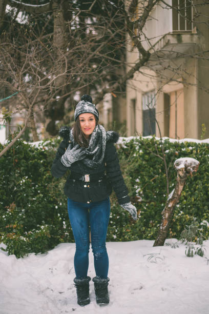 A woman standing outside at park in the snowfall. Happy female wearing a hat and scarf and cheerful smiling.