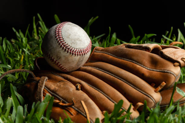 Studio shot of an old baseball resting on top of a catcher's mitt on grass with black background.
