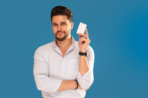 Handsome young bearded man holding credit card in hand, looking at the camera. Studio shot. Blue background. Copy space. Online payment.