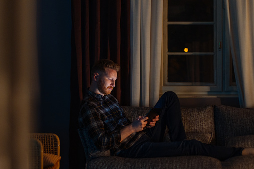 A side view of a pensive Caucasian freelancer casually dressed watching something online while sitting on the sofa in the living room at night.