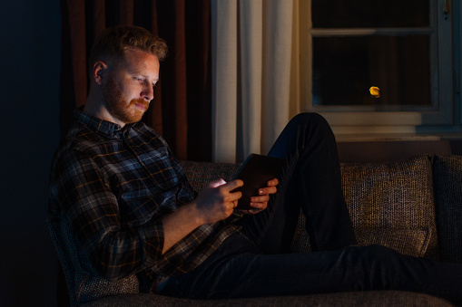A side view of a pensive Caucasian freelancer casually dressed watching something on his tablet while sitting on the sofa in the living room at night.