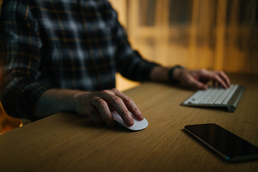 A cropped photo of an anonymous Caucasian businessman's hand holding a wireless mouse while reading something. He is sitting at the desk having his mobile phone nearby.