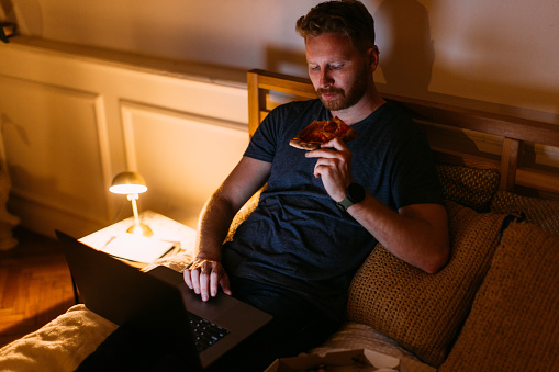 A serious Caucasian influencer scrolling on his laptop while lying in bed eating pizza.