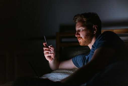 A pensive Caucasian influencer using his smartphone while lying in bed at night.