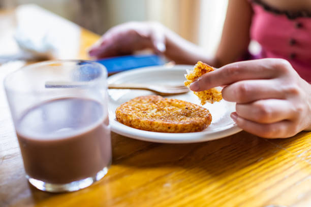 primo piano macro della donna seduta al tavolo della colazione che tiene mangiare patate fritte hash browns con mano sul piatto da vetro di latte al cioccolato e sfondo del telefono - 16330 foto e immagini stock