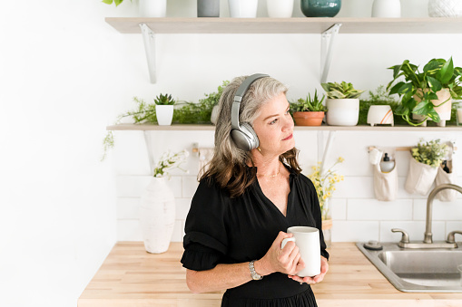 Woman listening to headphones in kitchen with plants holding mug