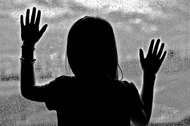 Little girl standing by window with raindrops on it on a rainy day