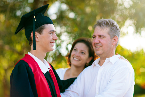 School or college graduation ceremony. Young man in gown and cap, with his family. Moher and father cheer at celebration of successful diploma certificate. High school graduate in robe and mortarboard