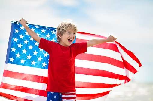 Kids run with USA flag on sunny beach. 4th of July celebration. American family fun on Independence Day weekend. Patriotic children celebrate US holiday. Boy and girl with symbols of America.