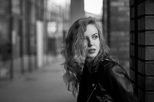 attractive curly blond girl in leather jacket standing on city street looking at camera over shoulder, monochrome