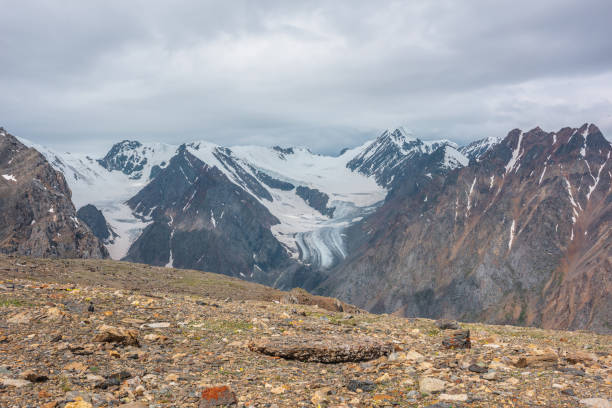 istock Dramatic landscape with high snowy mountain range with long glacier tongue and pointed peak in sunlight under cloudy sky. Atmospheric view from stone hill to large snow mountains at changeable weather 1404762733